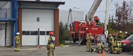 strażacy podczas ćwiczeń na placu przed JRG, w tle samochody osobowe, słupki ostrzegawcze i rozstawiony podnośnik