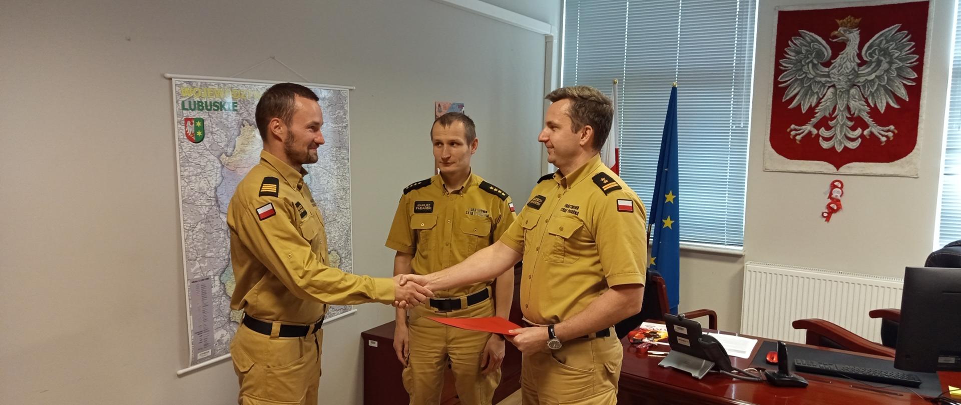 Fotografia przedstawia Komendanta Powiatowego Państwowej Straży Pożarnej w Strzelcach Krajeńskich wręczającego w gabinecie decyzję o przyznaniu nagrody funkcjonariuszowi z Jednostki Ratowniczo-Gaśniczej PSP w Strzelcach Krajeńskich.