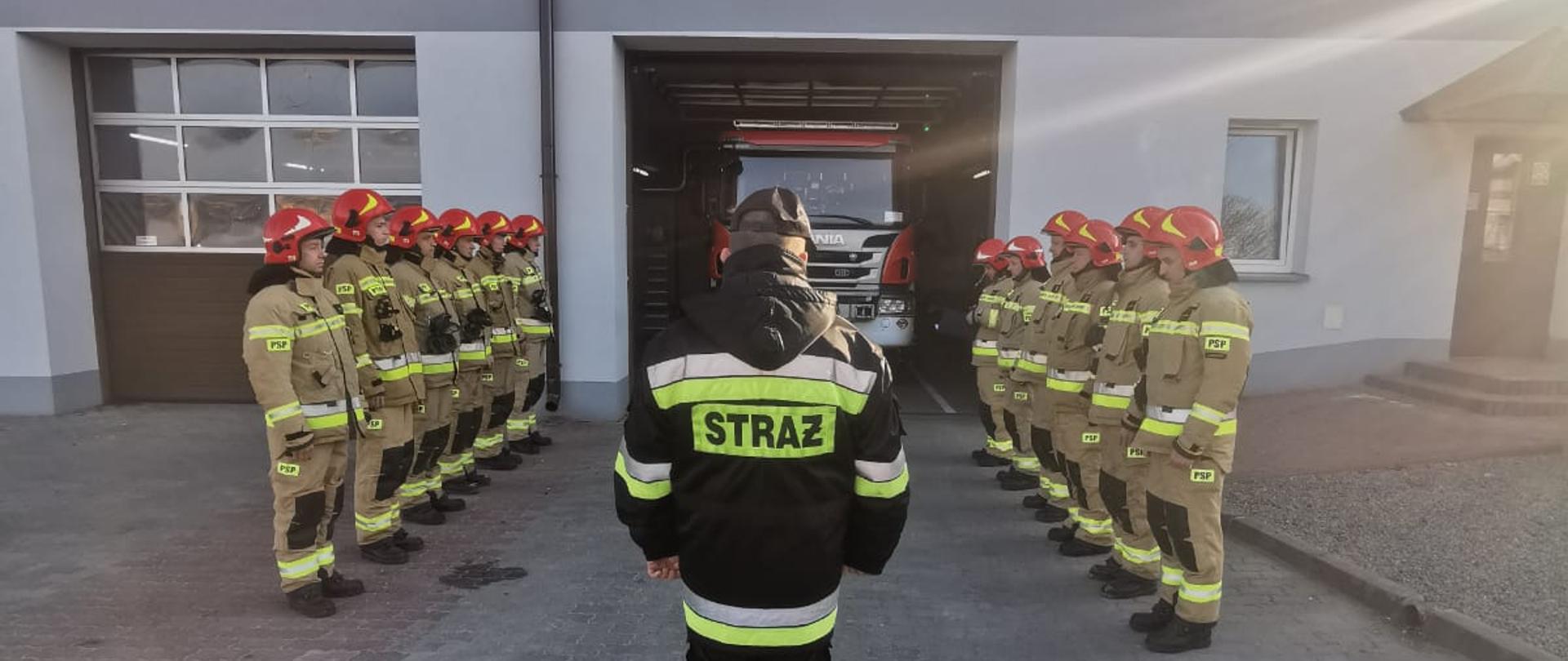 Zdjęcie przestawia strażaków stojących w dwuszeregu przed remizą PSP.
