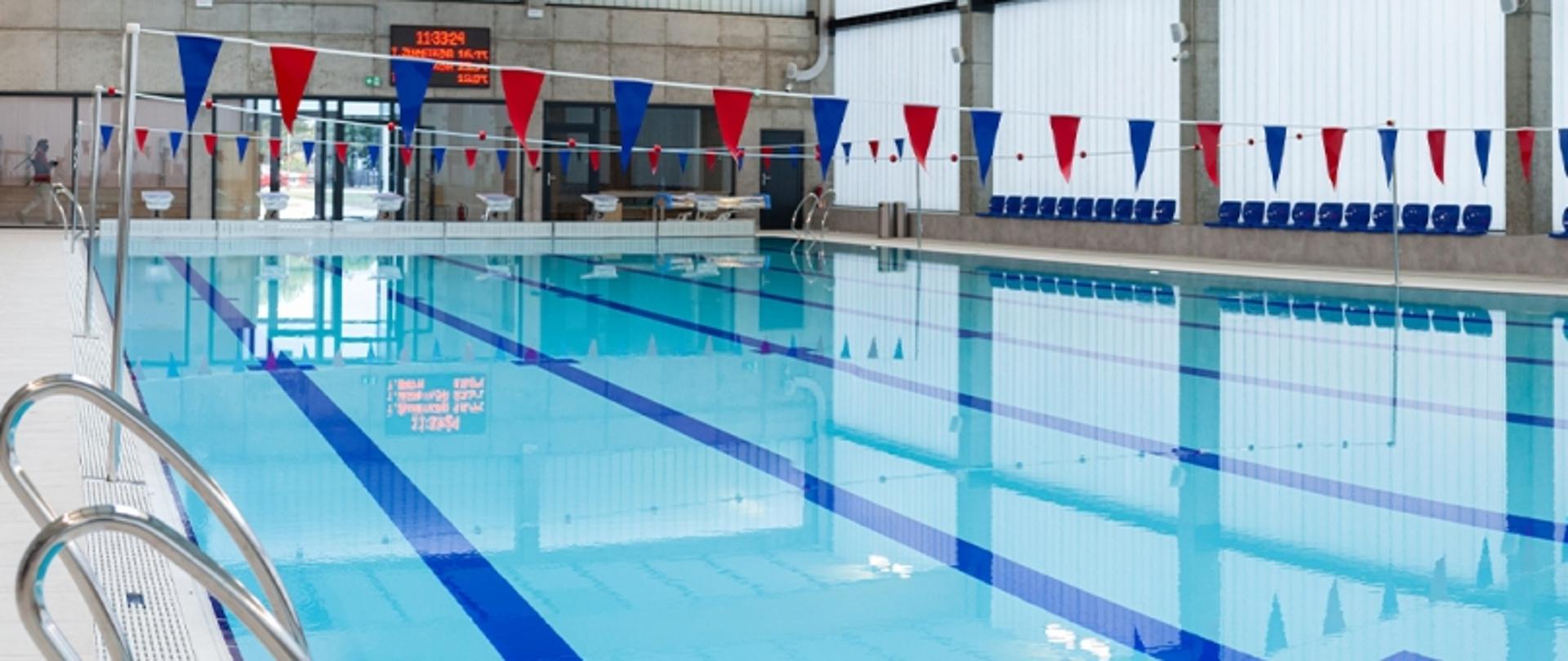 Kryty basen z torami pływackimi, chorągiewkami nad wodą i niebieskimi krzesłami przy ścianie; w tle zegar cyfrowy.