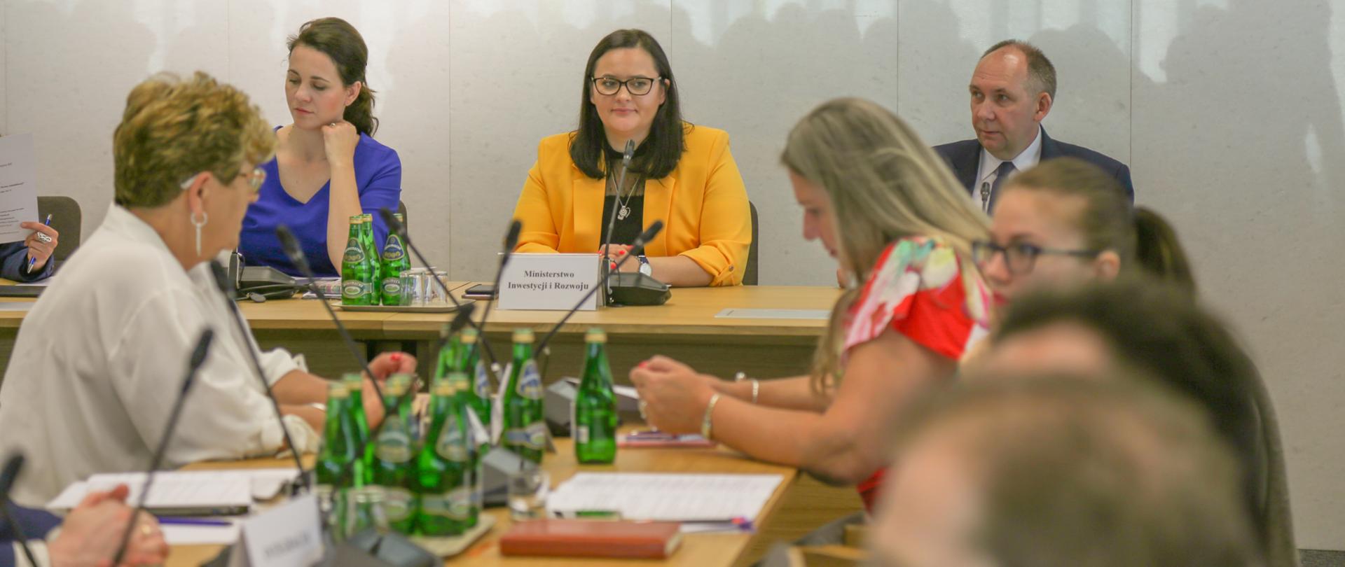 Grupa ludzi siedzi na sali. Przy stole prezydialnym siedzą trzy osoby. Po środku wiceminister Małgorzata Jarosińska-Jedynak.