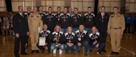 Sportowcy ustawieni w dwuszeregu z medalami na piersiach. Obok zawodników stoją strażacy w umundurowaniu. 