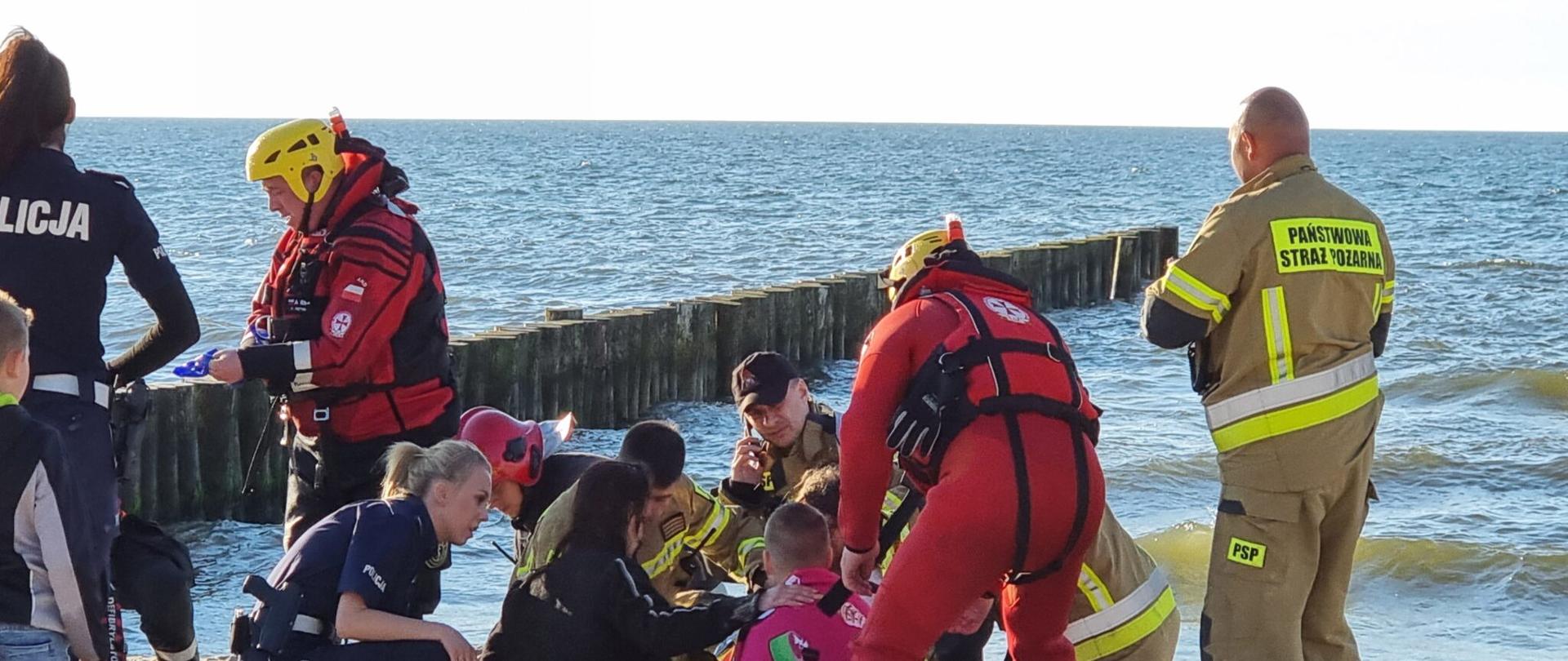 Zdjęcie przedstawia akcję ratunkową na plaży.Widać pracowników różnych służb, w tle widać morze.