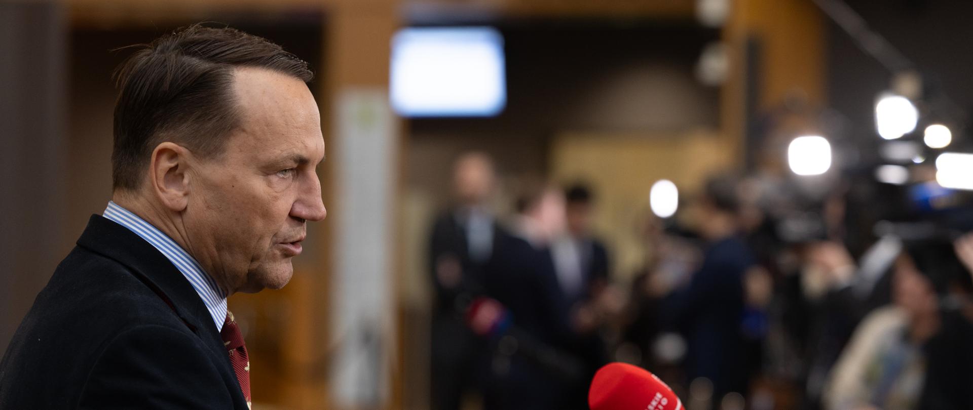  Radosław Sikorski w garniturze stoi przy mikrofonie. 