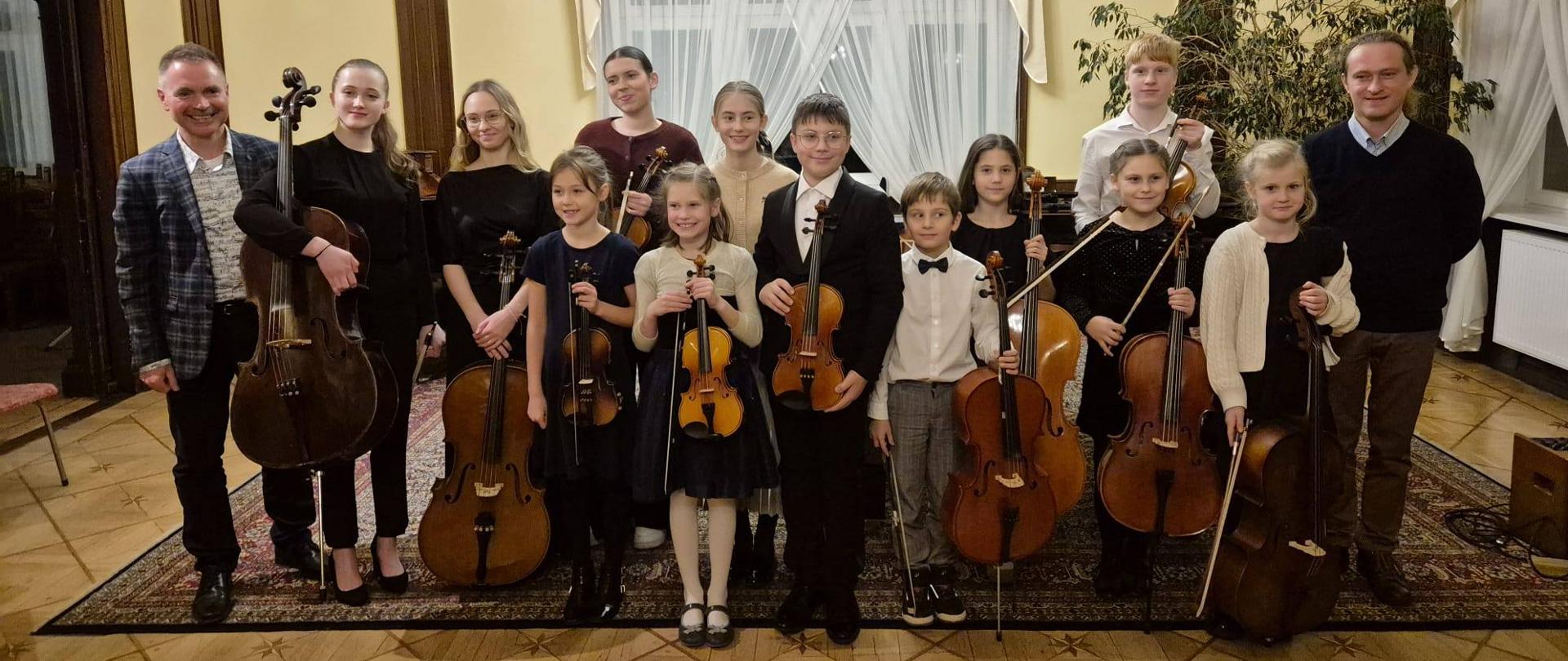 Zdjęcie kolorowe, wykonane w eleganckim wnętrzu. Grupa dwunastu osób, w tym dwóch mężczyzn i dziesięciu młodych osób, stojąca na tle jasnego okna. Dziewięć osób trzyma instrumenty strunowe: wiolonczele i skrzypce. Mężczyzna po lewej stronie w jasnej marynarce, mężczyzna po prawej w czarnym swetrze. Osoby pośrodku ustawione w rzędach. W tle żółte ściany, zasłonięte okno, żyrandol i wzorzysty dywan na drewnianej podłodze.