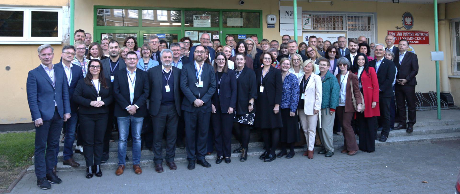 Duże zdjęcie grupowe osób (mężczyźni i kobiety) stojących przed budynkiem. Na szyldzie nad wejściem widnieje napis: "KSZTAŁCIMY 70 LAT DLA RZEMIOSŁA" oraz "BRANŻOWE CENTRUM UMIEJĘTNOŚCI".