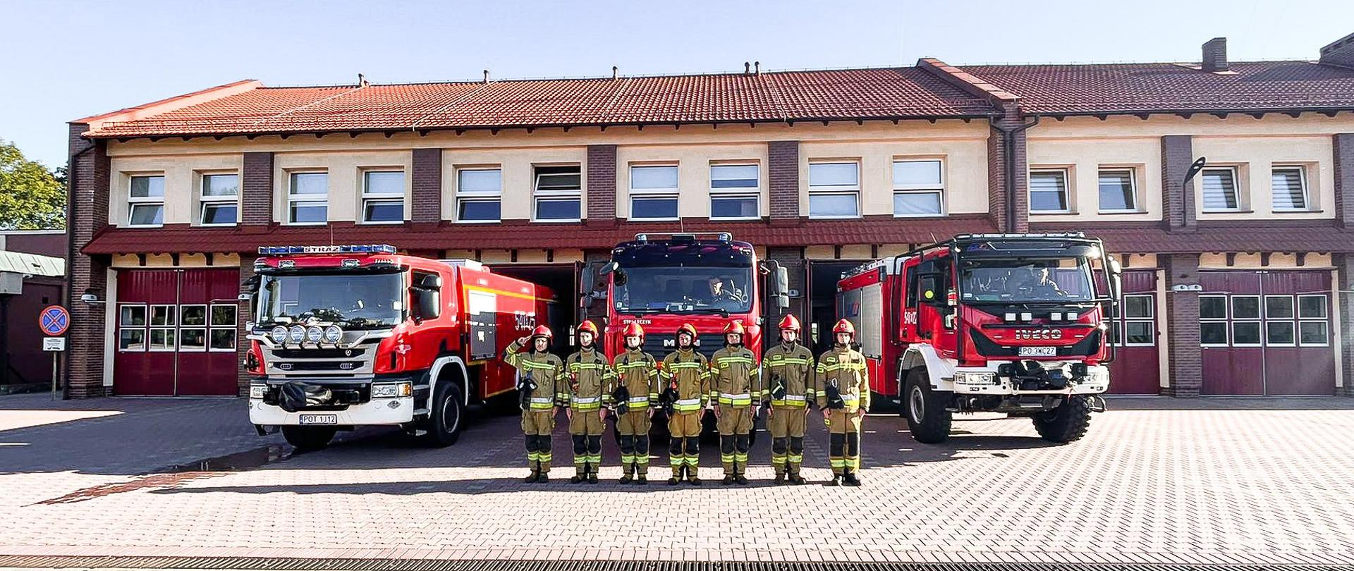 Strażacy w ubraniach specjalnych stoją przed wozami strażackimi i oddają hołd. W tle budynek strażnicy.
