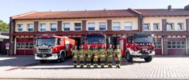 Strażacy w ubraniach specjalnych stoją przed wozami strażackimi i oddają hołd. W tle budynek strażnicy.