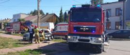 Zdjęcie przedstawia samochód po kolizji stojący na chodniku. Obok niego stoi samochód strażacki w włączonymi sygnałami świetlnymi. Przy rozbitym samochodzie stoją strażacy, a za nimi ratownicy medyczni z lekarzem i karetką oraz dwa kolejne pojazdy pożarnicze.