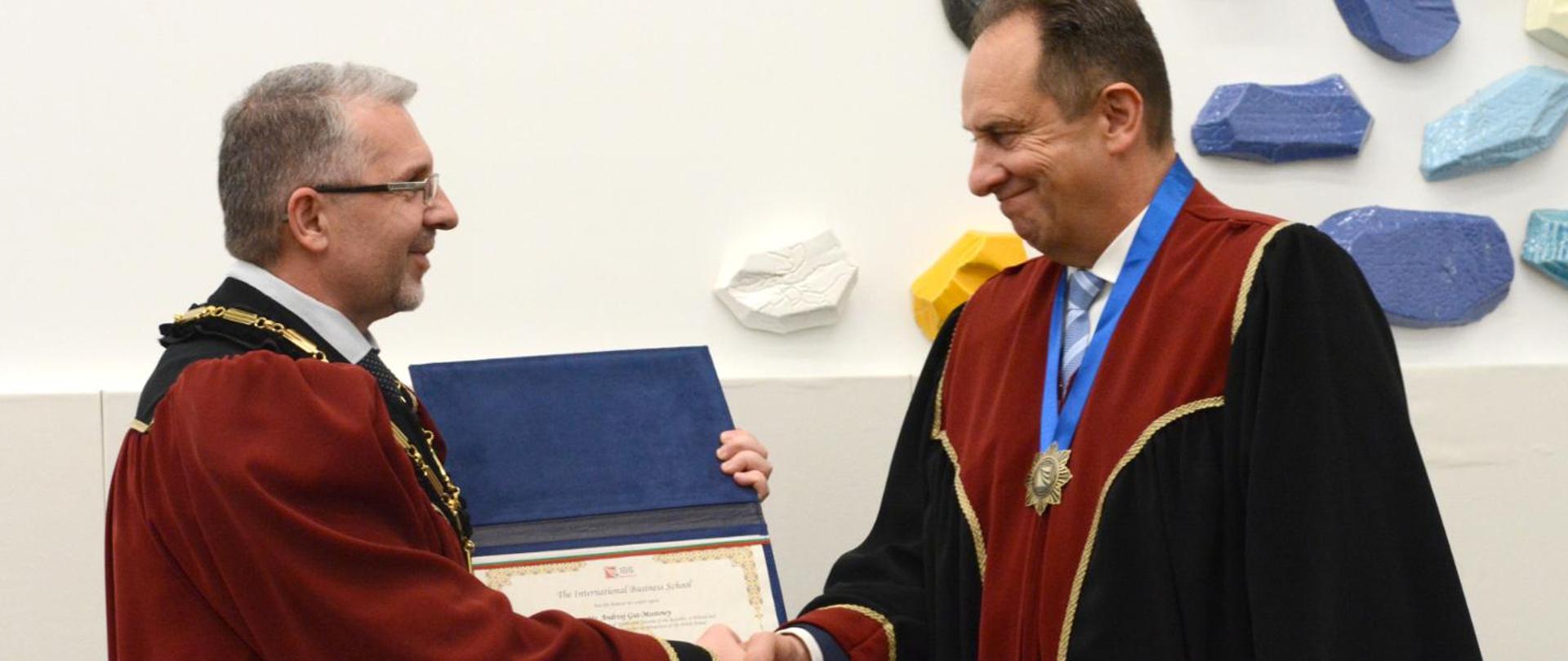 Two men in togas shaking hands. Andrzej Gut-Mostowy wearing a medal and receiving a diploma.