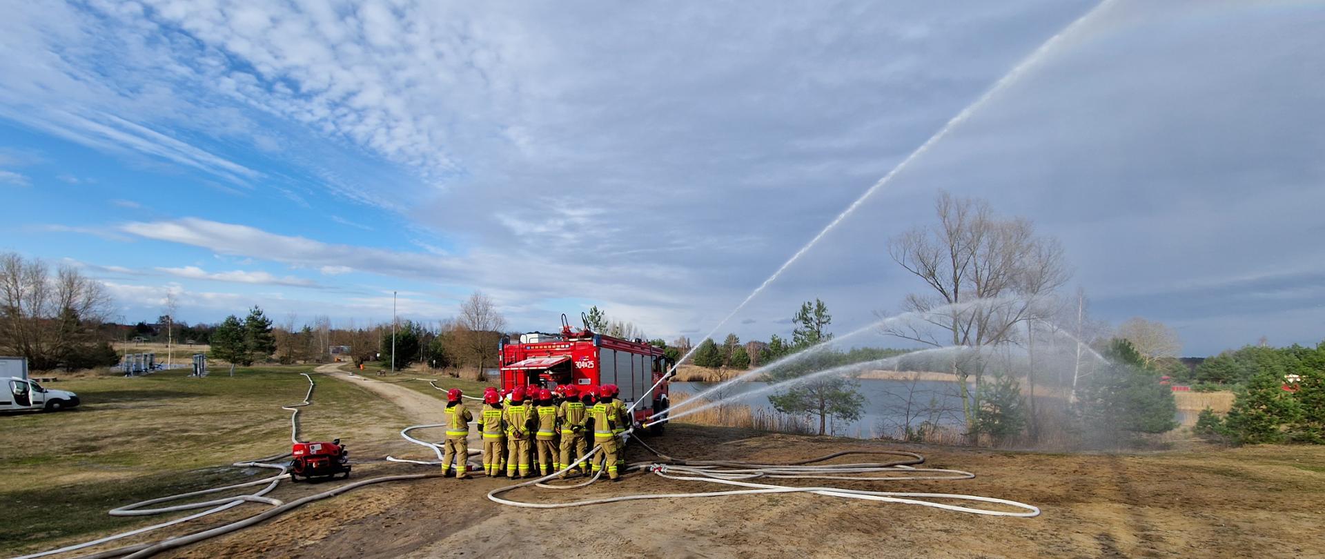 strażacy stoją przy samochodzie, wokoło rozwinięte węże z wodą