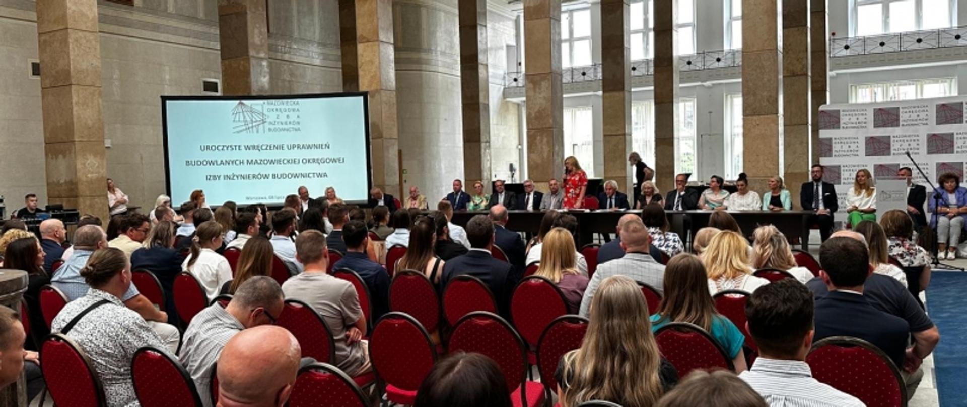 Sala z uczestnikami wydarzenia siedzącymi w czerwonych krzesłach, scena z prelegentami i ekranem z napisem o uroczystym wręczeniu uprawnień budowlanych.