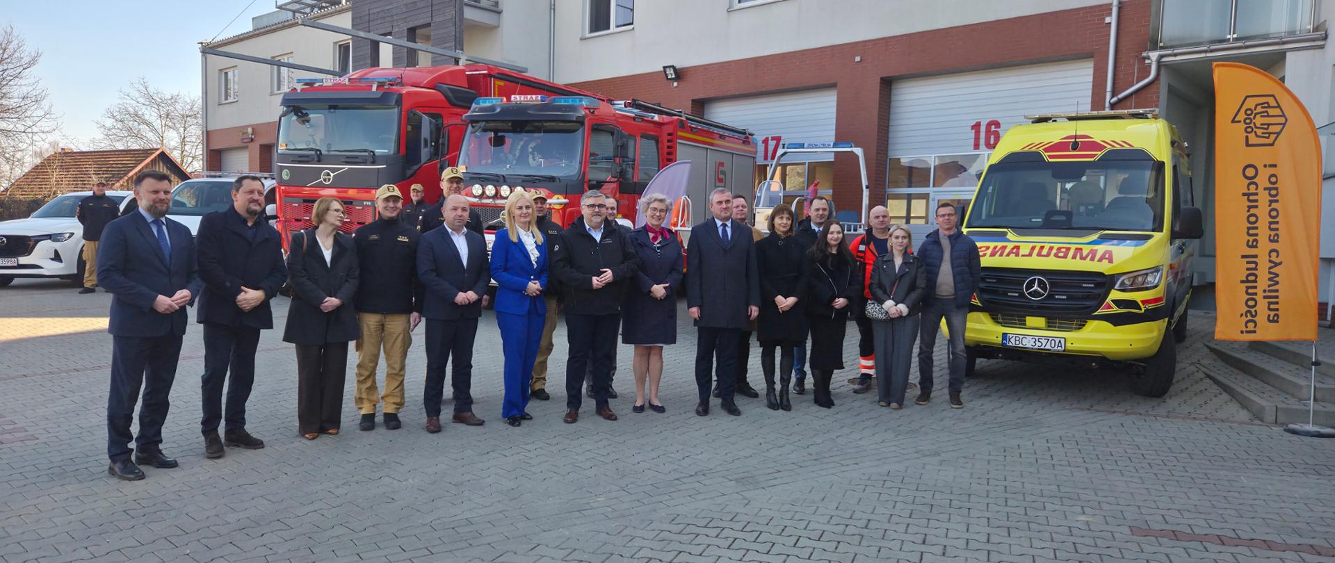 Fotografia przedstawia grupę zaproszonych gości oraz funkcjonariuszy stojących w szeregu na placu przed komendą. Na pierwszym planie wyróżnia się kobieta w niebieskim garniturze oraz mężczyźni w eleganckich płaszczach i garniturach. Obok nich stoją strażacy w piaskowych mundurach służbowych. Wszyscy z powagą uczestniczą w oficjalnej części uroczystości, a w tle widać fragmenty zaparkowanych wozów bojowych.