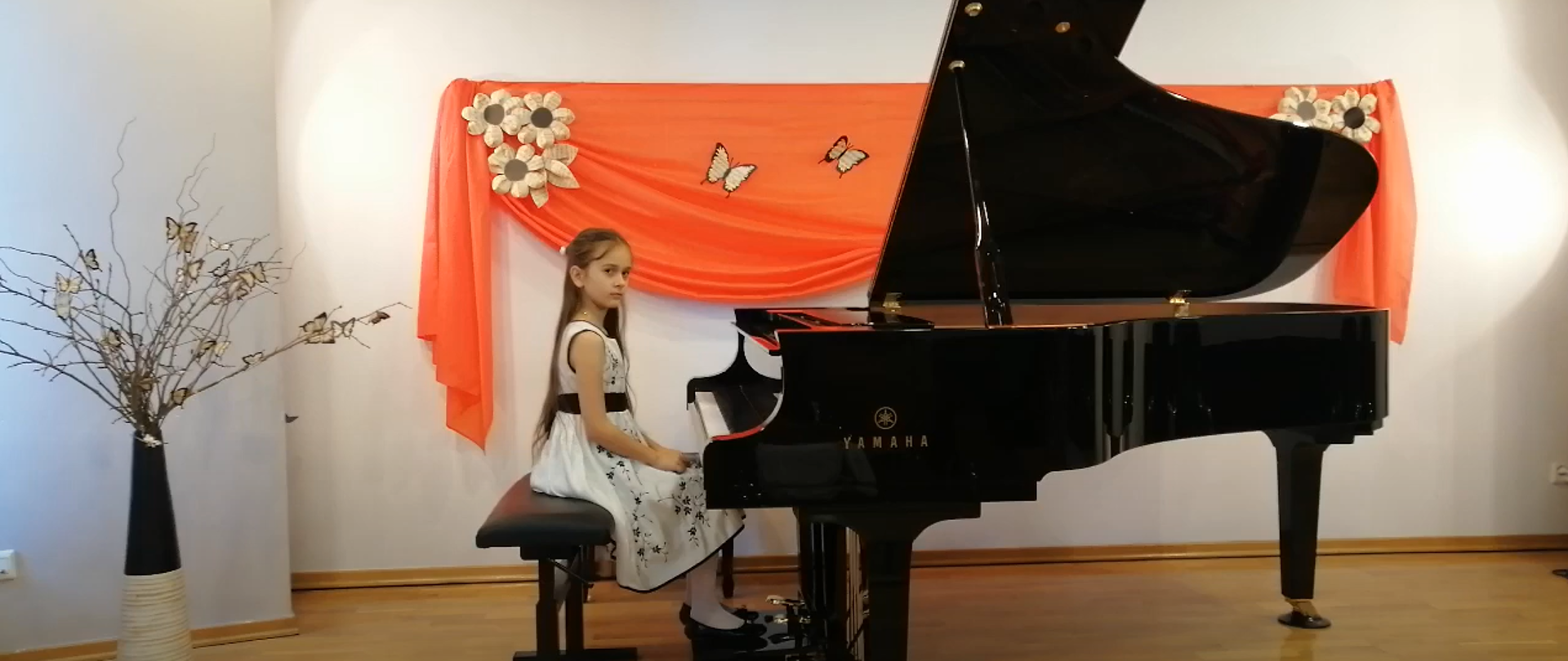 Na zdjęciu uczennica szkoły siedząca przy fortepianie. W tle dekoracja składająca się z pomarańczowego materiału,, na którym umocowano beżowe kwiatki oraz motylki.Z lewej strony zdjęcia wazon, a w nim gałęzie i beżowe motylki.