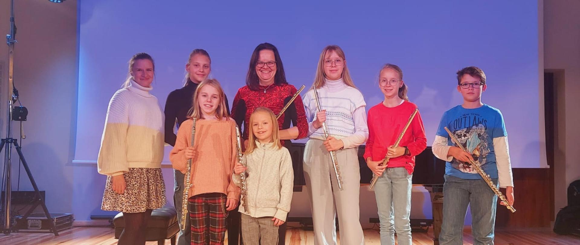 Zdjęcie przedstawia uczniów klasy fletu stojących na deskach sceny wraz z nauczycielem panią Kingą Waszak i osobą prowadzącą warsztaty fletowe dr Alicją Molitorys. Nauczyciel stoi jako pierwszy od lewej, natomiast osoba prowadząca na środku za uczniem. Uczniowie trzymają w dłoniach flety. W tle jest wysunięty ekran z rzuconym niebieskim światłem. Wszyscy ubrani są na kolorowo. 