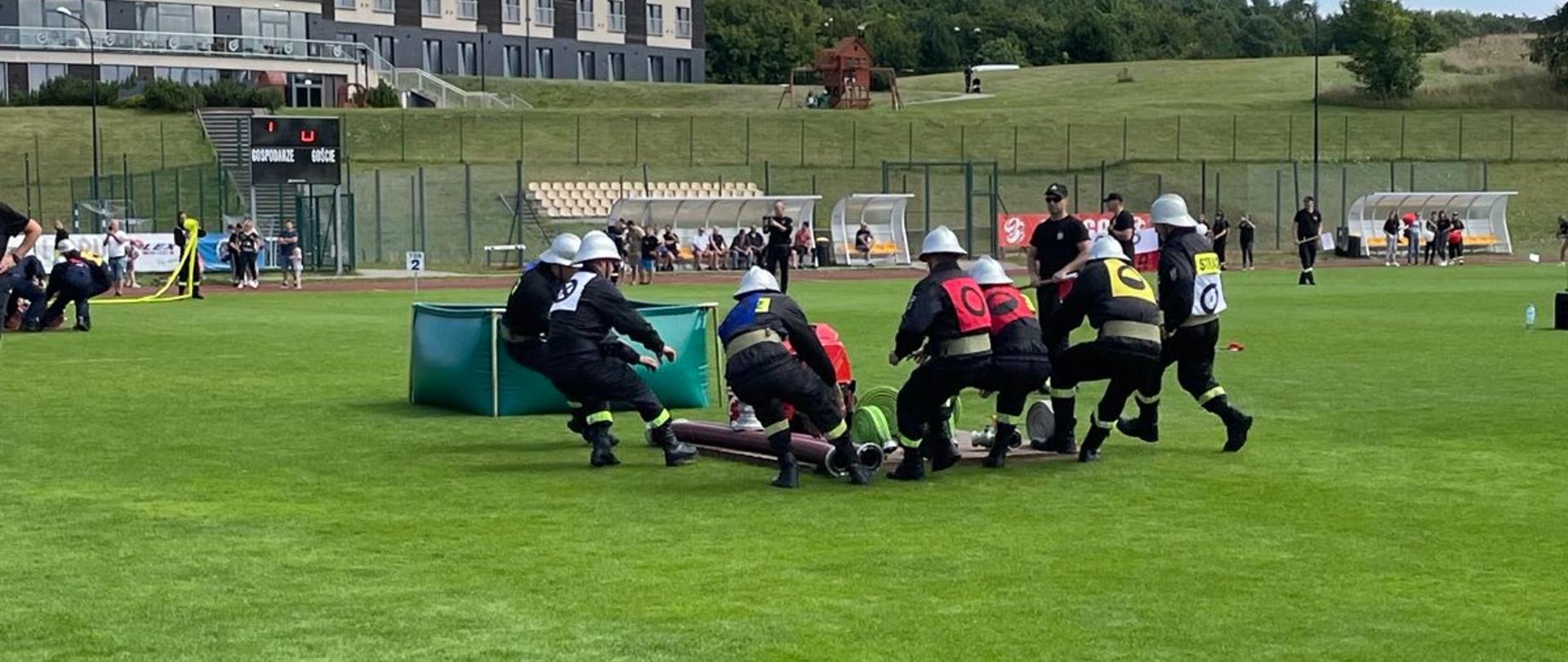 Teren boiska trawiastego. Drużyna strażaków ujęta w biegu do podestu ze sprzętem pożarniczym podczas startu w ćwiczeniu bojowym, obok zbiornik z wodą, z tyłu kibice.