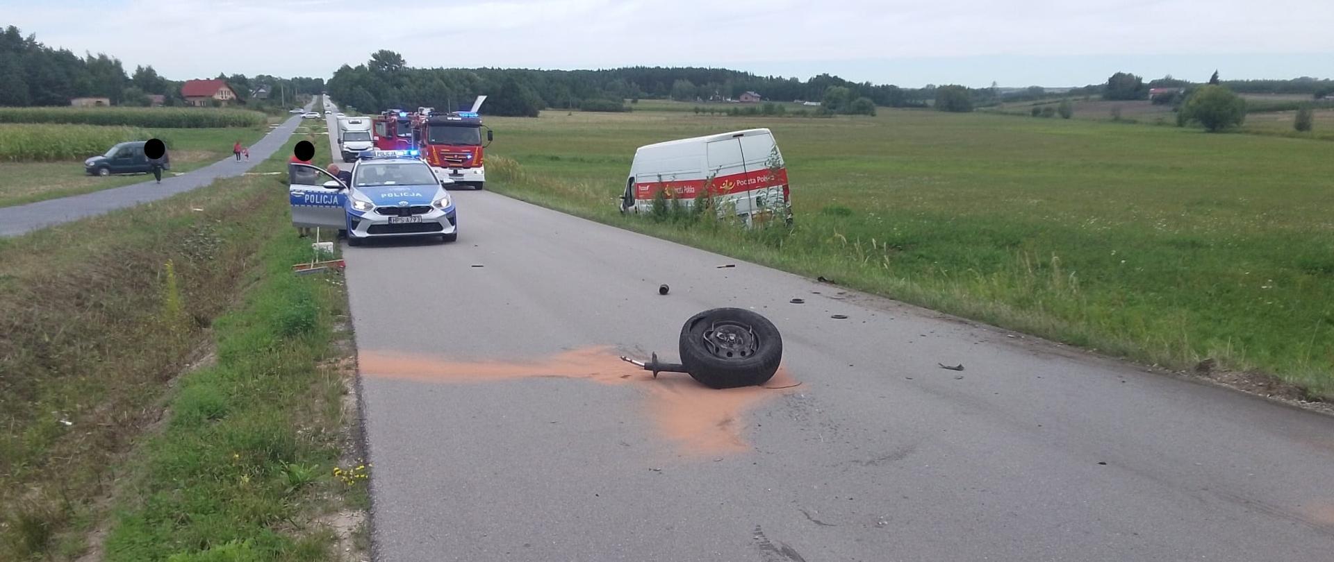 Na drodze: Asfalt jest usiany licznymi, drobnymi szczątkami powypadkowymi.
Po lewej: Stoi karetka pogotowia (ambulans) na sygnale, a za nią wóz strażacki.
Po prawej: W rowie i na poboczu znajduje się mocno zniszczony, ciemnoszary samochód osobowy z otwartą maską i uszkodzonym przodem.