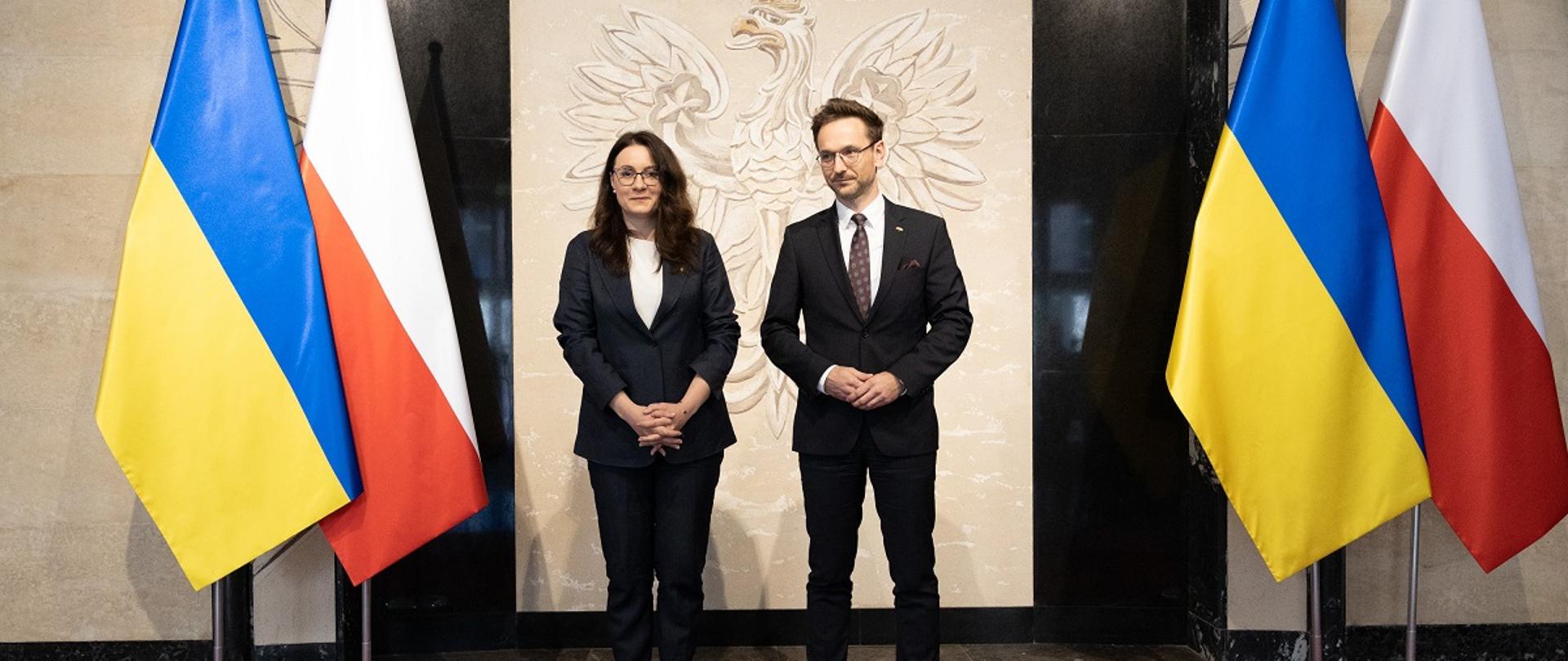 Minister rozwoju i technologii Waldemar Buda oraz wicepremier i minister gospodarki Ukrainy Julia Svyrydenko stoją na tle flag Polski i Ukrainy, za ich plecami orzeł w koronie.