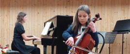 Dziewczynka siedząc na krześle na środku estrady gra na wiolonczeli, za nią na pianinie gra kobieta.