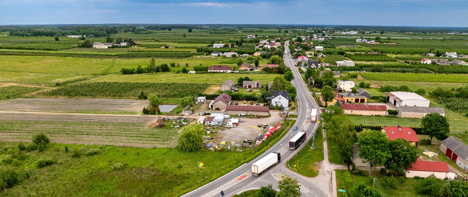 Widok z góry na jednojezdniową drogę przebiegającą przez niewielką miejscowość. Po drodze jedzie kilka samochodów ciężarowych. Dookoła dużo zieleni: łąki, drzewa, zarośla