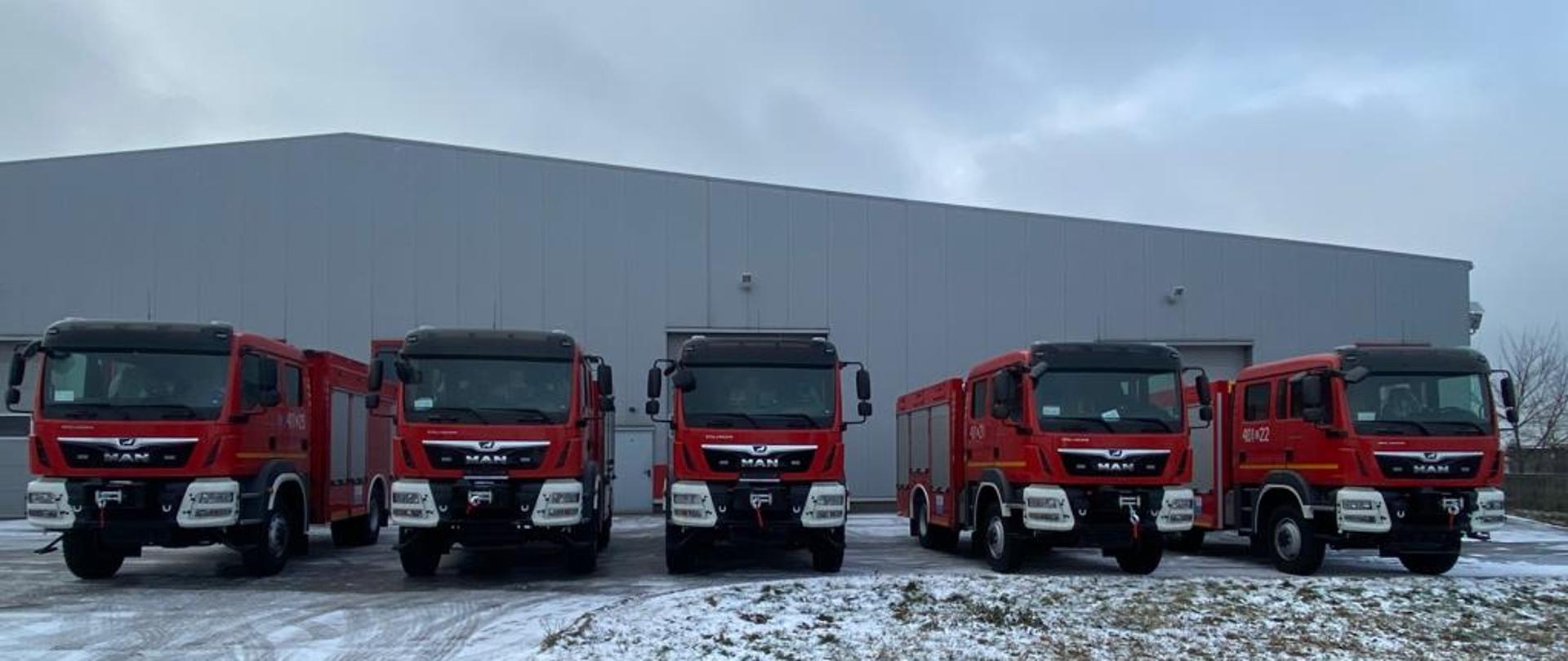 Na zdjęciu widać pięć samochodów ratowniczo gaśniczych ustawionych obok siebie. Zdjęcie zrobione z przodu pojazdów. Pojazdy stoją na zaśnieżonym placu przed halą wykonawcy pojazdów.