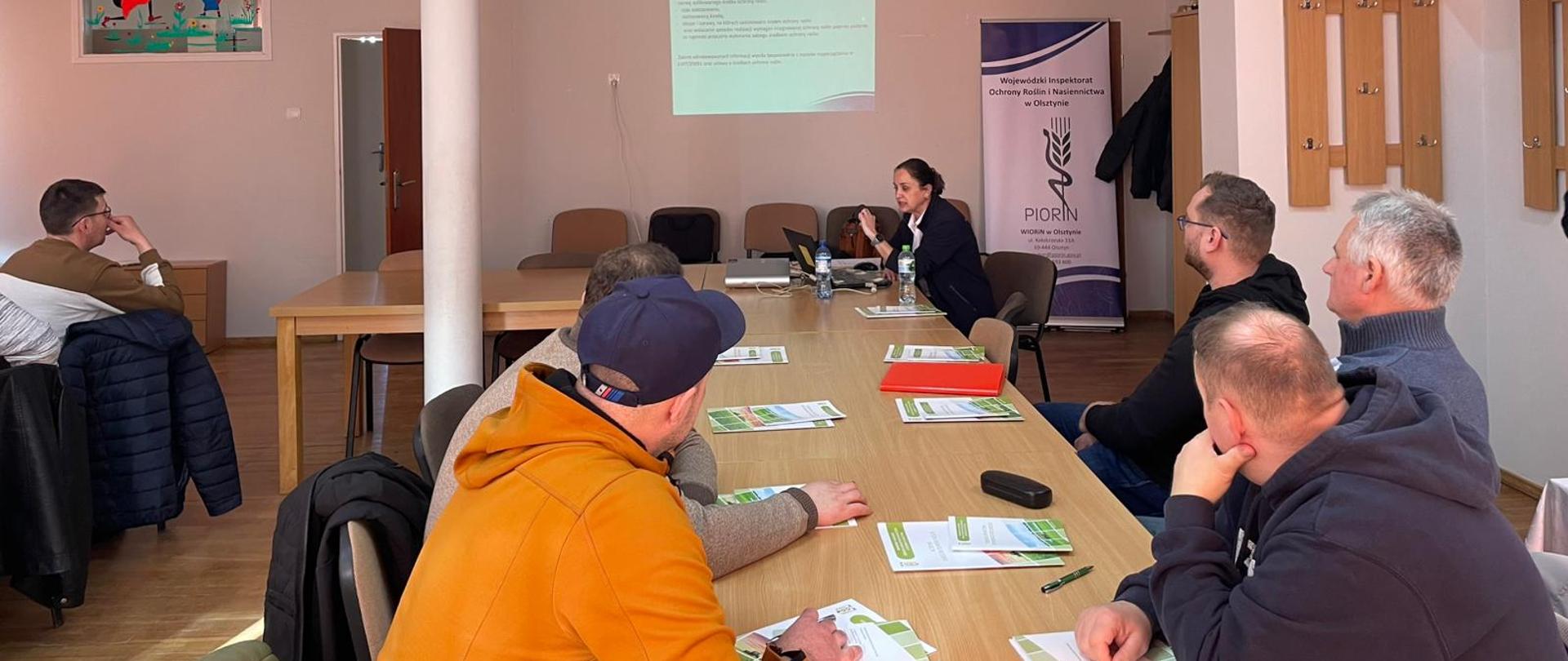 grupa osób siedzi przy stołach zwróceni do kobiety gestykulującej w głębi zdjęcia, za plecami ma rollup PIORiN