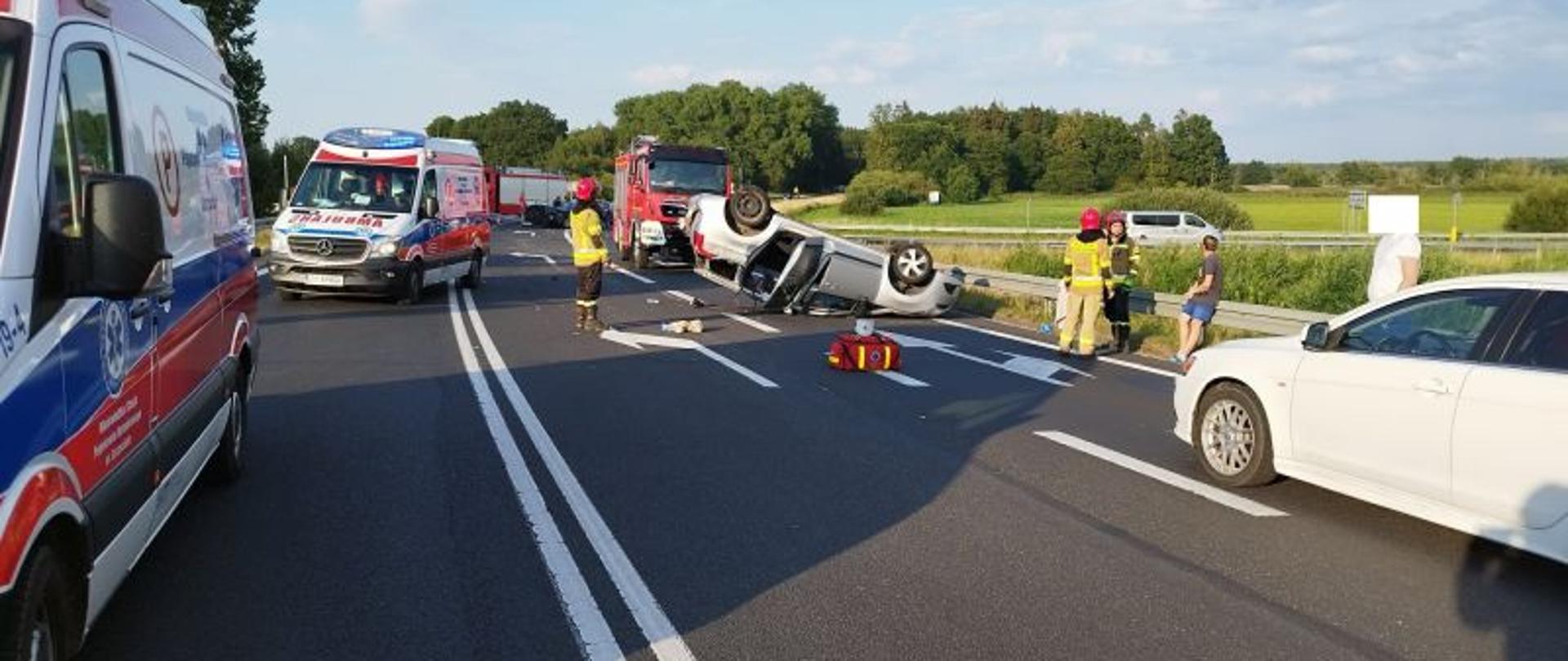 Zdjęcie przedstawia miejsce zdarzenia. Widać na nim pojazdy uczestniczące w zdarzeniu, strażaków i inne służby. Jedno z aut leży na dachu.