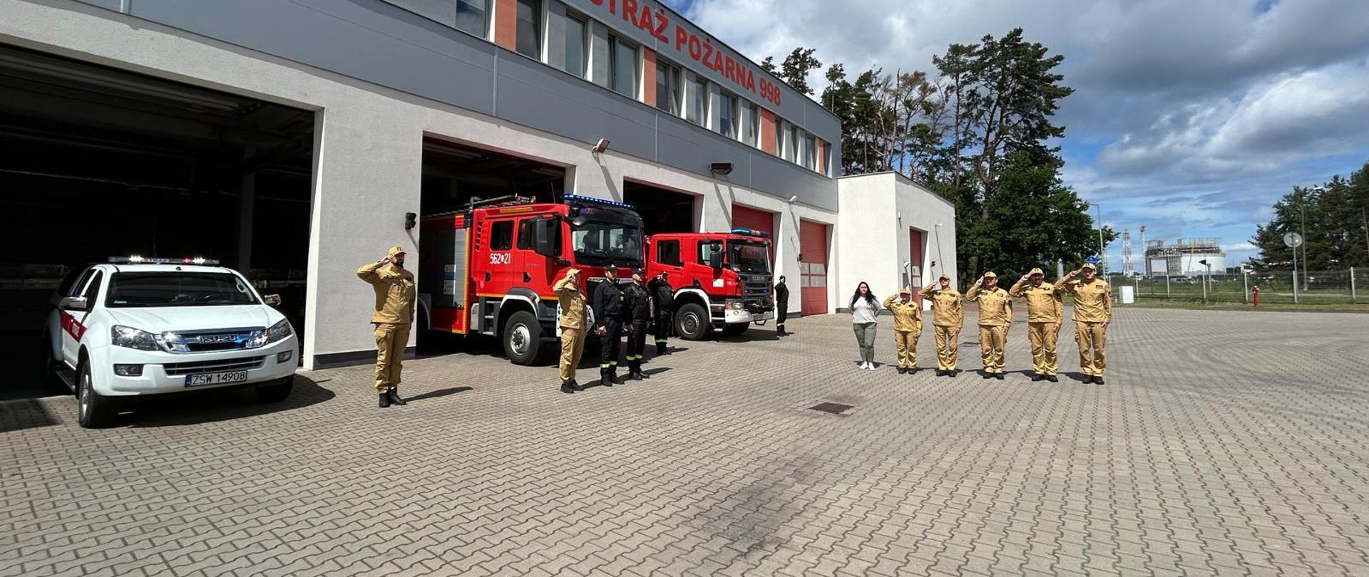 Na zdjęciu widoczni funkcjonariusze KM PSP w Świnoujściu stojący na kostce brukowej i oddający honor. W tle budynek komendy oraz samochody pożarnicze. Od lewej samochód osobowy koloru białego, następnie dwa pojazdy ciężarowe koloru czerwonego. Elewacja budynku szara. 