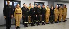 Grupa strażaków w mundurach służbowych — czarnych i piaskowych — stoi w równych szeregach wewnątrz jasnej sali. Po lewej stronie stoi oficer w mundurze galowym. Wszyscy patrzą przed siebie w formalnej postawie jak podczas uroczystości.

