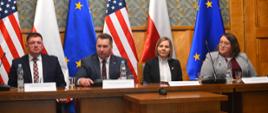 Przy stole siedzą od lewej wiceminister Rzymkowski, minister Czarnek, dyrektor ORPEG Justyna Kralisz oraz kobieta ubraną w szarą marynarkę. W tle flagi: polskie, amerykańskie i unijne. 