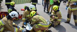 Na ziemi leżą dwa manekiny umieszczone na deskach ortopedycznych. Strażacy znajdujący się w pozycji klęczącej udzielają kwalifikowanej pierwszej pomocy. Wokół strażaków znajdują się torby z wyposażeniem służącym do udzielania kwalifikowanej pierwszej pomocy. Dwóch strażaków stoi i sporządza karty z informacją z udzielonej pomocy. Dwóch strażaków trzyma koc w celu zasłonięcia miejsca działań. W tle znajdują się obserwatorzy w kamizelkach.
