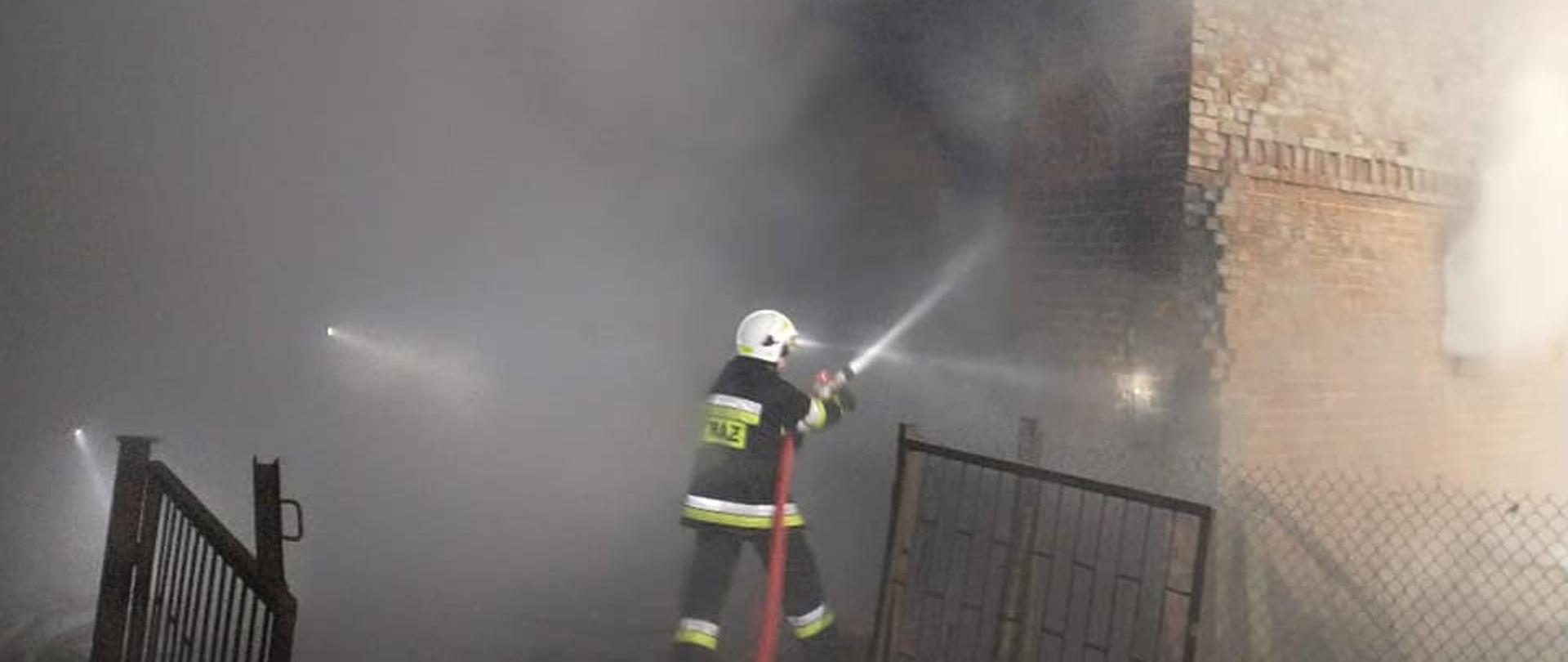 Zdjęcie przedstawia strażaka OSP prowadzącego działania gaśnicze na budynek inwentarski, z którego wydobywa się dym