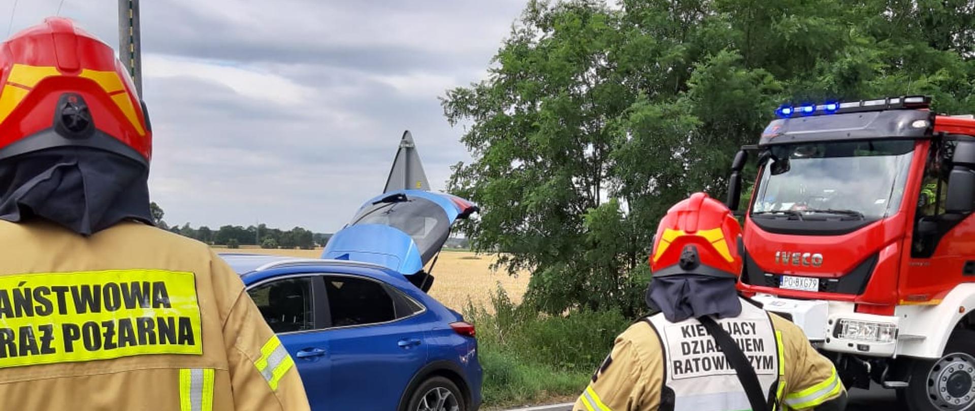 na zdjęciu strażak w kamizelce kierującego działaniem ratowniczym na jezdni posypany sorbent obok stoi samochód ratowniczo - gaśniczy i osobowy niebieski z otwartą tylną klapą obok strażaka stoi drugi strażak z radiostacją 