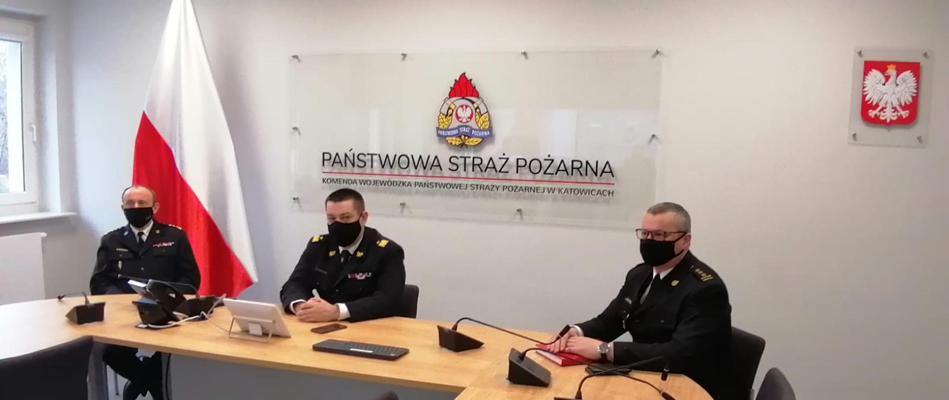 Zdjęcie przedstawia wideokonferencję na sali sztabowej Komendy Wojewódzkiej PSP w Katowicach. Przy stole konferencyjnym, w środku siedzi Śląski Komendant Wojewódzki PSP, po jego lewej i prawej stronie zastępcy. Komendanci ubrani są w mundury wyjściowe, na twarzach mają maseczki ochronne.