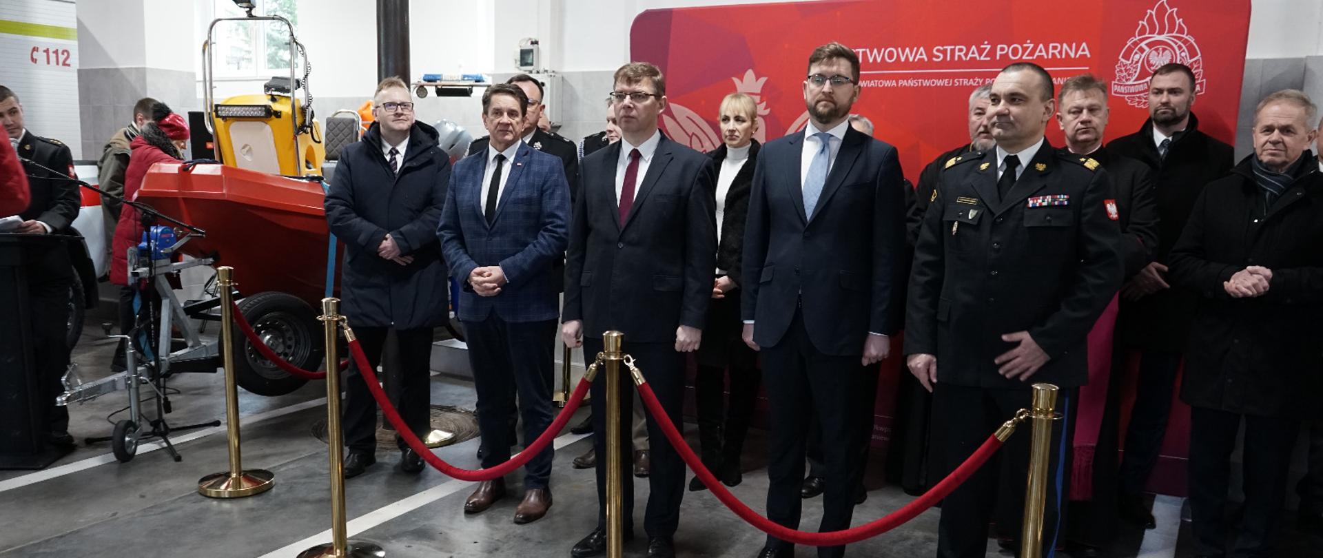 Garaż bojowy strażaków. Na tle białej ścian i czerwonego banera stoją zaproszeni goście. Z lewej strony widoczny przód łodzi motorowej.