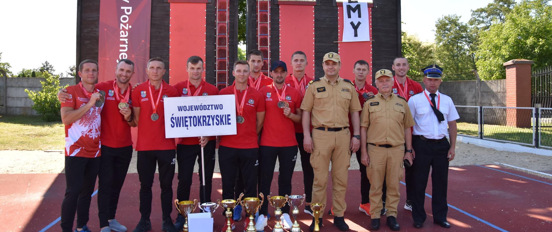 Zdjęcie przedstawia drużynę reprezentującą województwo świętokrzyskie na 38 Mistrzostwach Polski w Sporcie Pożarniczym. Zawodnicy w czerwonych koszulkach stoją na tle wspinali. Na szyjach mają medale różnych kolorów. Na ziemi stoją okazałe puchary. Z zawodnikami jest także świętokrzyski komendant wojewódzki nadbryg. Krzysztof Ciosek wraz z zastępcą- st. bryg. Jarosławem piotrowskim.