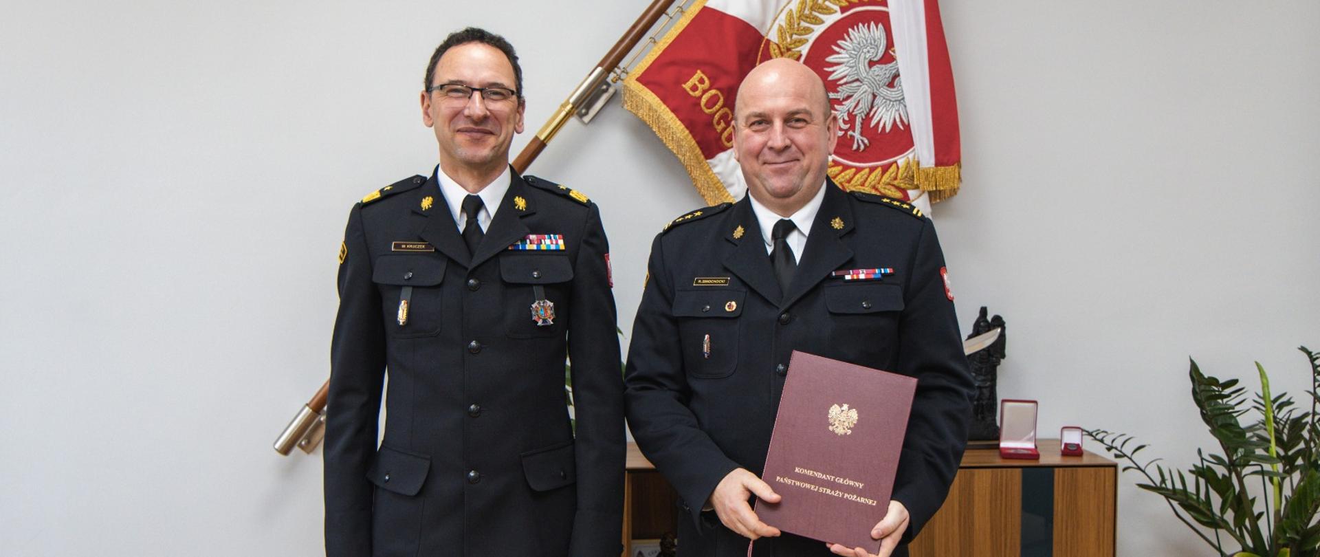 Dwóch umundurowanych oficerów Państwowej Straży Pożarnej stoi obok siebie w pomieszczeniu biurowym. Oficer po prawej stronie trzyma bordowy dokument z godłem Rzeczypospolitej Polskiej. W tle widoczna jest flaga PSP oraz szafka z pamiątkami.