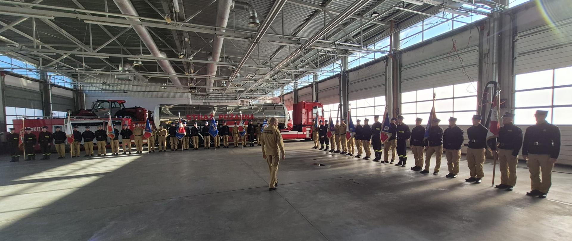 Strażacy PSP w piaskowych mundurach służbowych z czarnymi polarami oraz w czarnych mundurach koszarowych stoją w szeregu tworząc literę L po dwóch stronach garażu. Za nimi stoi srebrno-czerwona cysterna z napisem „STRAŻ POŻARNA”. W rękach trzymają sztandary.
