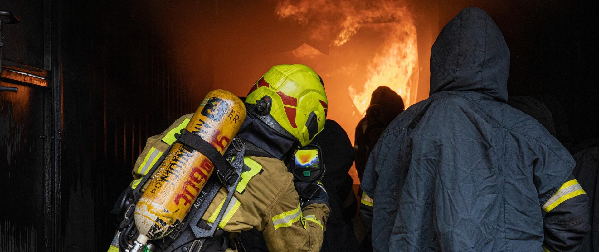 Strażacy podczas ćwiczeń w komorze rozgorzeniowej. W tle płomienie.