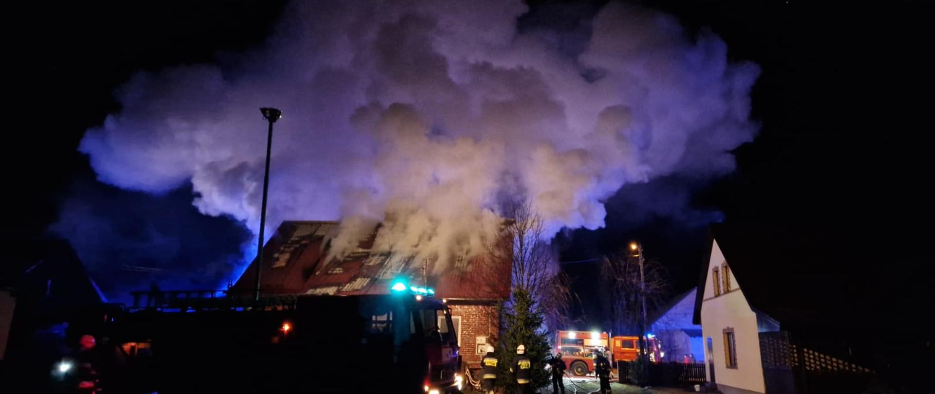 Zdjęcie przedstawia zadymienie nad domem, który objęty został pożarem, w tle jednostki straży pożarnej