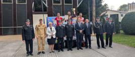 W dwóch rzędach stoją funkcjonariusze Państwowej Straży Pożarnej, oraz zaproszenie oficjele w pierwszym rzędzie, w drugim rzędzie na podium stoją sportowcy, na szyjach mają zwieszone medale, a w rękach dyplomy. Z tyłu widać wspinalnię.