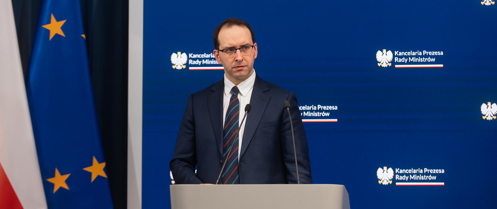 Minister Stanisław Żaryn na tle granatowego tła z logo KPRM podczas konferencji prasowej