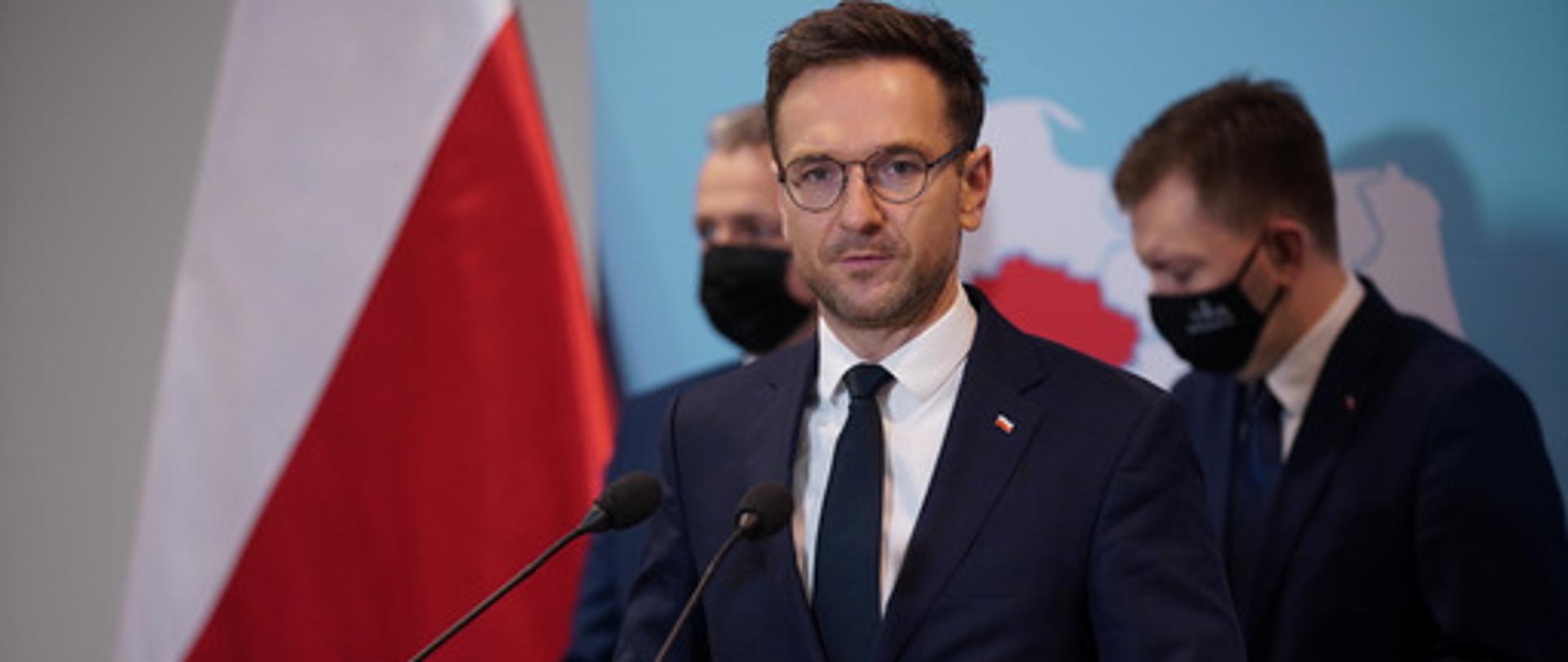 W mównicy wiceminister Waldemar Buda. Za nim dwóch mężczyzna w maseczkach. Obok flaga Polski.