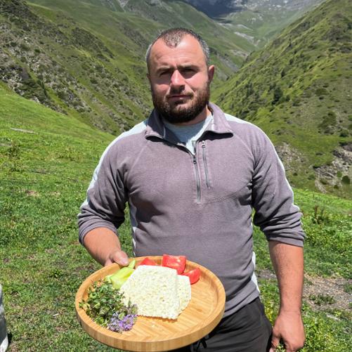 Na tle gór stoi mężczyzna, który trzyma w ręku drewnianą tacę z serem, warzywami i ziołami