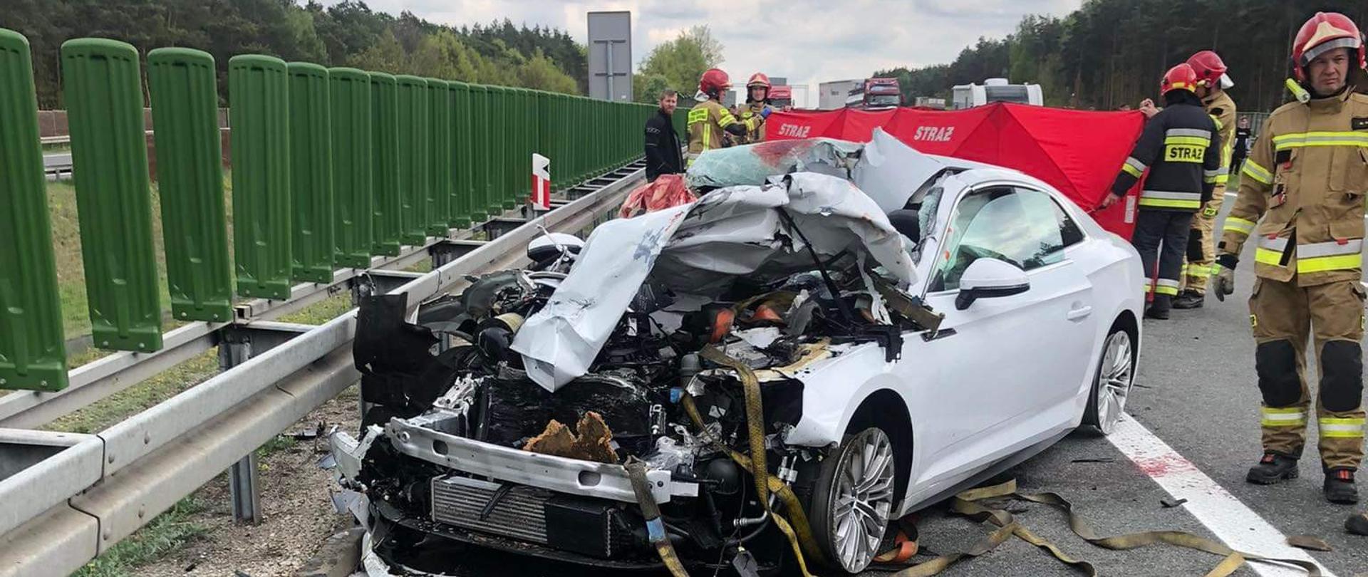 Zdjęcie przedstawia rozbity samochód osobowy koloru białego przy barierach energochłonnych. Po lewej stronie znajdują się zielone osłony przeciwolśnieniowe. Z pozostałych storn rozbitego pojazdu widać stojących, ubranych na żółto strażaków oraz resztki powypadkowe porozrzucane na jezdni. W tle widnieją fragmenty czerwonego parawanu strażackiego, lasu oraz zachmurzonego nieba.