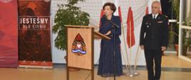 Wręczenie promes. Sala sportowa w Szkaradowie. Przy mównicy przemawia do zebranych Pani minister Marlena Maląg. Z przodu na mównicy umieszczony jest logotyp PSP.. Obok przemawiającej stoi strażak PSP w umundurowaniu wyjściowym. W tle flagi państwowe, kwiat oraz materiały promujące KP PSP w Rawiczu