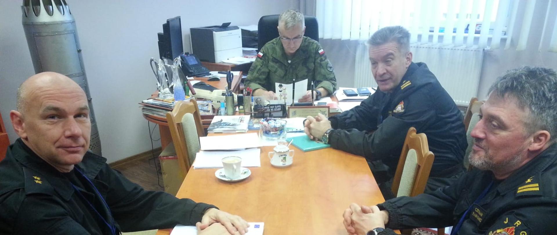 Zdjęcie przedstawia spotkanie czterech mężczyzn. Siedzą oni w gabinecie przy stołach. Trzech z nich jest strażakami w czarnych mundurach, zaś czwarty jest wojskowym w mundurze moro. Na biurku znajdują się dokumenty oraz różne przybory.