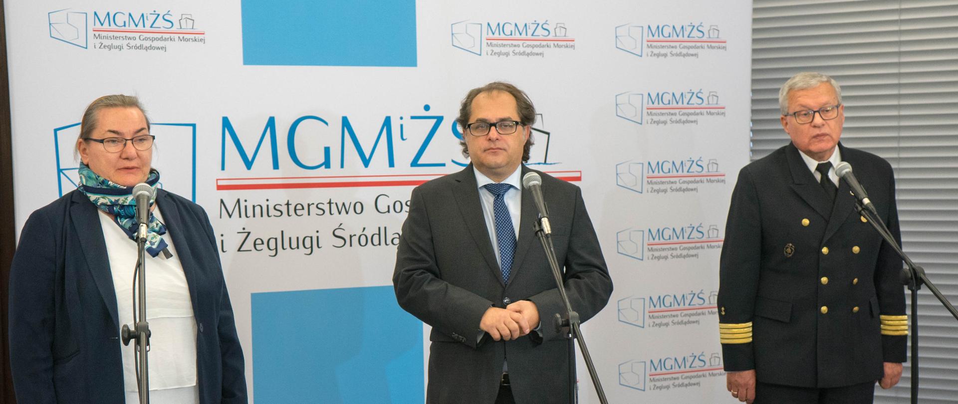Zdjęcie przedstawia prelegentów konferencji na tle loga MGMiŻŚ. Minister Marek Gróbarczyk (po środku), dyrektor Urzędu Morskiego w Gdyni Wiesław Piotrzkowski (po prawej) oraz zastępca dyrektora Urzędu Morskiego ds. technicznych Anna Stelmaszyk–Świerczyńska (po lewej).
