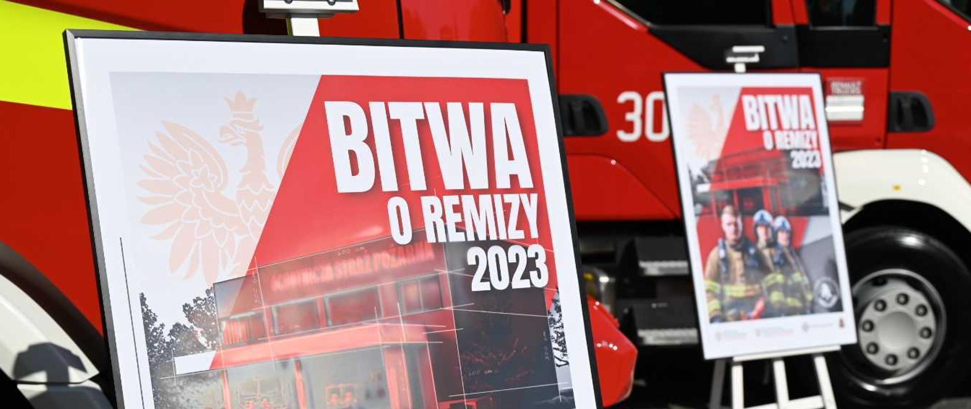 Zdjęcie przedstawia plakaty akcji "Bitwa o remizy 2023", które stoją przed wystawionymi samochodami pożarniczymi.