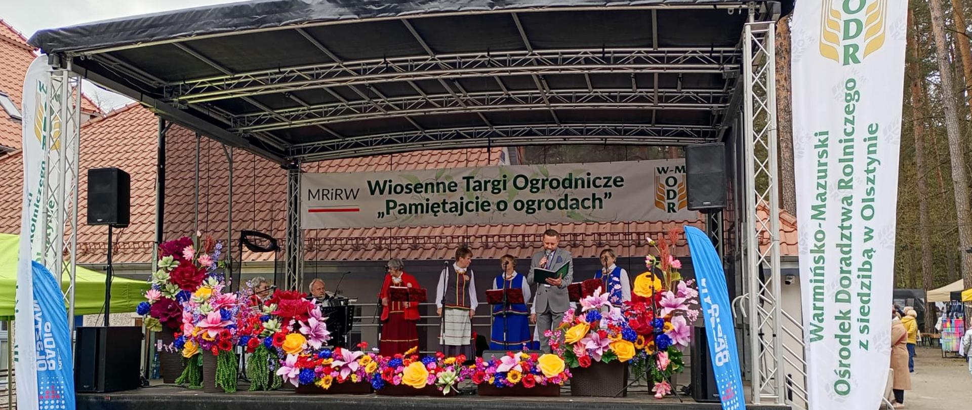 na scenie z prawej strony stoi mężczyzna i czyta tekst do mikrofonu, za nim grupa ludzi w ludowych strojach. dookoła banery 